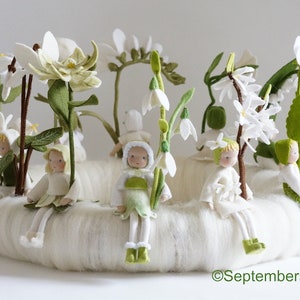 Op de afbeelding: Een handgemaakte vilten bloemenkrans met miniatuurfiguren. De krans heeft witte bloemen, groene bladeren en kleine poppen gekleed in wit en groen. De poppen zitten rond de krans en creëren een grillige scène.