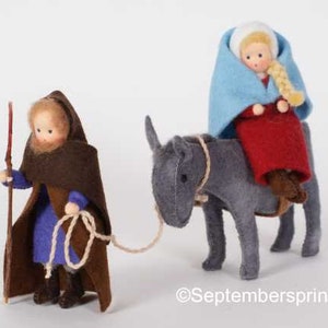 May include: A felt doll dressed in a brown robe and blue tunic holds a stick. A second felt doll dressed in a blue and red robe sits on a gray donkey. The donkey is being led by a rope held by the first doll.