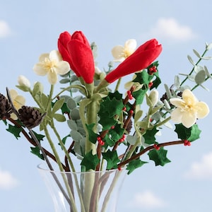 May include: A bouquet of artificial flowers in a clear glass vase featuring bright red tulip-like blooms, cream-colored blossoms, green holly leaves with small red berries, brown pinecones, and light green eucalyptus-style foliage stems.