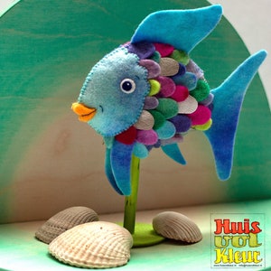 Puede incluir: Un pez de fieltro azul con escamas de colores y una boca amarilla. El pez está de pie sobre una base de madera con conchas a su alrededor. El texto "Huis vol Kleur" es visible en la esquina inferior derecha de la imagen.