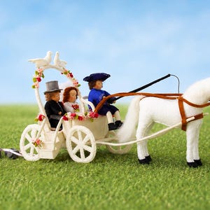 May include: A miniature white horse-drawn carriage featuring a driver in a blue coat and two passengers in formal wedding attire. The carriage is decorated with pink and red flowers and two white doves on top, with small gray bells trailing behind.