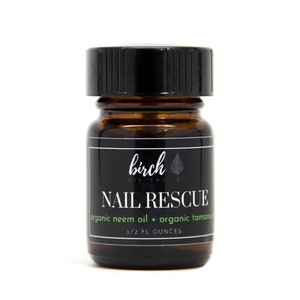 May include: A small, brown glass bottle with a black lid. The label reads "NAIL RESCUE" with "organic neem oil + organic tamanu" and "1/2 FL OUNCES" below. The brand name "birch sisters" is at the top of the label.