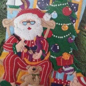 May include: A felt Christmas stocking kit featuring Santa Claus in red pajamas, a Christmas tree, and wrapped gifts. Santa is holding a toy and sitting in a rocking chair. A teddy bear sits on a drum. The background includes a window and a sailboat.