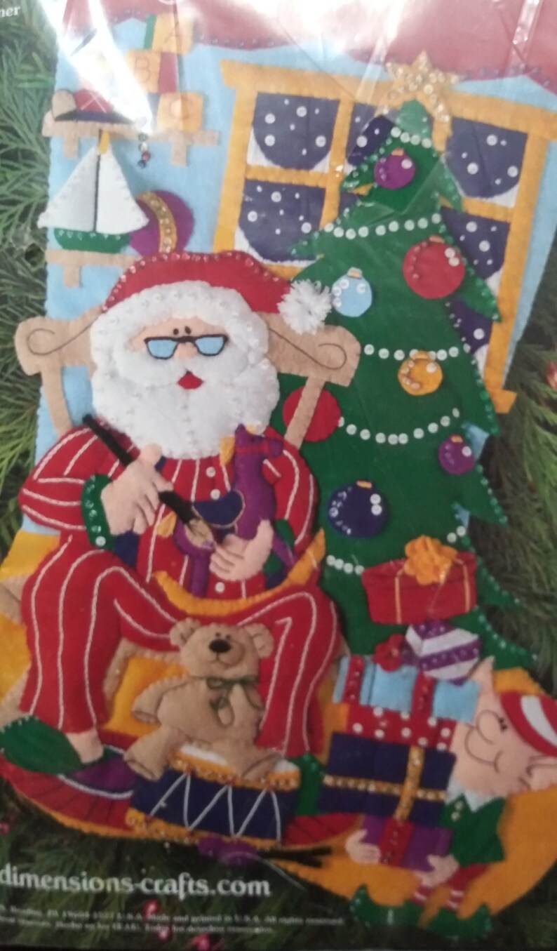 May include: A felt Christmas stocking kit featuring Santa Claus in red pajamas, a Christmas tree, and wrapped gifts. Santa is holding a toy and sitting in a rocking chair. A teddy bear sits on a drum. The background includes a window and a sailboat.