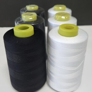 May include: A collection of six cylindrical spools of sewing thread arranged in two rows. Three spools are black and three are white. Each spool features a bright yellow plastic core at the top, and some are wrapped in clear protective plastic film.