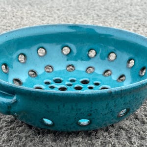 May include: A turquoise ceramic colander with numerous circular drainage holes. The bowl has a rounded shape with a small handle on one side. The glazed surface reflects light, highlighting the color and texture of the pottery.