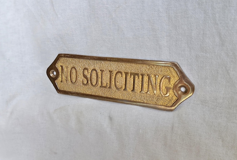 May include: A rectangular brass sign with pointed ends featuring the embossed text NO SOLICITING in capital letters. The background of the sign has a textured finish, while the border and lettering are smooth and polished. Two screw holes are visible.