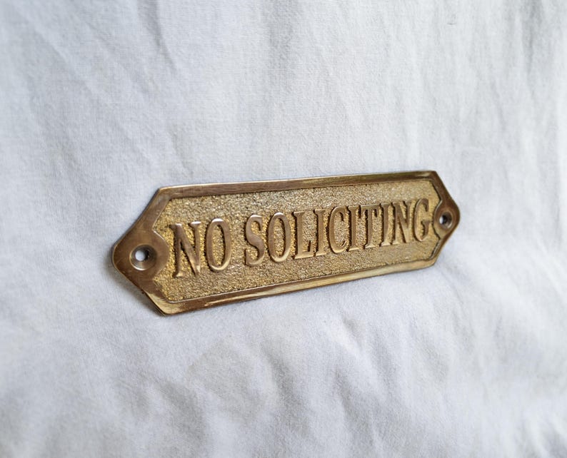 May include: A rectangular brass sign with beveled edges and pointed ends, featuring the embossed text NO SOLICITING in capital letters. The background of the text has a textured finish, while the border and letters are smooth and polished.