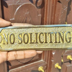 May include: A rectangular brass sign with beveled edges and a textured gold-tone background. The sign features the raised, smooth-finished text "NO SOLICITING" in capital letters. There is a circular mounting hole on each of the two pointed ends.