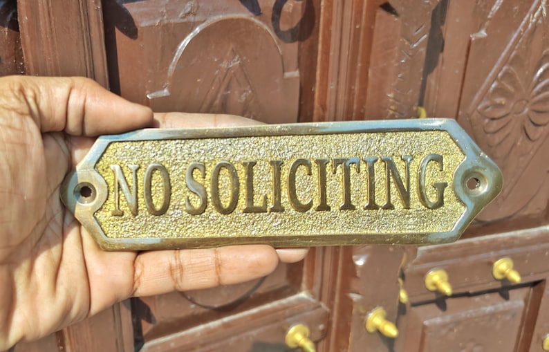 May include: A rectangular brass sign with beveled edges and a textured gold-tone background. The sign features the raised, smooth-finished text "NO SOLICITING" in capital letters. There is a circular mounting hole on each of the two pointed ends.