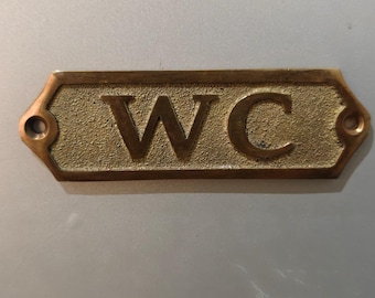May include: A rectangular brass sign with beveled edges and two mounting holes on the sides. The sign features a textured background with the letters WC embossed in a smooth, raised font. The metal has a warm, metallic gold-toned finish.