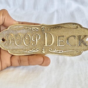 Könnte beinhalten: Ein goldfarbenes Metallschild mit strukturierter Oberfläche und geschwungenen Rändern. Der Text POOP DECK ist mittig in einer Serifenschrift eingeprägt. Kleine Ankersymbole befinden sich über und unter dem Text, sowie zwei Schraublöcher.