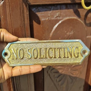 May include: A rectangular brass sign with beveled edges and a textured gold-tone center featuring the embossed text NO SOLICITING in capital letters. The sign includes two pre-drilled mounting holes on the left and right sides for installation.