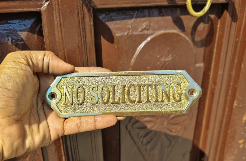 May include: A rectangular brass sign with beveled edges and a textured gold-tone center featuring the embossed text NO SOLICITING in capital letters. The sign includes two pre-drilled mounting holes on the left and right sides for installation.