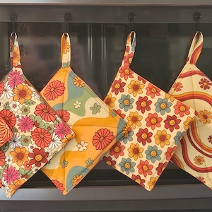 May include: A set of six square pot holders with hanging loops, each featuring a different retro floral or groovy design in shades of orange, yellow, pink, and blue. The pot holders are hanging on a dark metal bar.