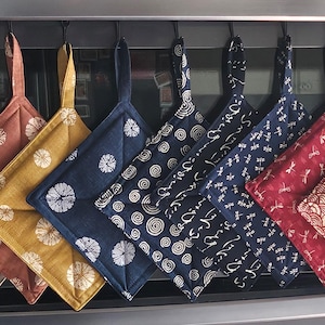 Puede incluir: Una colección de nueve agarradores de tela cuadrados colgados en fila. Los agarradores presentan varios patrones, incluyendo círculos, espirales, libélulas y motivos de ondas en colores beige, óxido, mostaza, azul marino y rojo con detalles blancos.