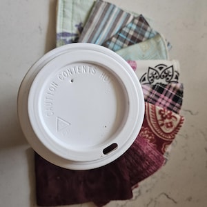 May include: A white plastic coffee cup lid with the text CAUTION CONTENTS HOT embossed on the top, positioned over a fan of various fabric coffee cup sleeves featuring assorted patterns including stripes, plaid, paisley, and floral designs.