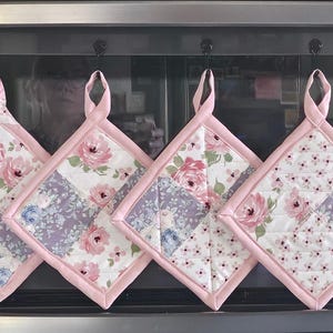 Peut inclure: Quatre maniques carrées matelassées suspendues en rangée, chacune présentant un motif patchwork de roses roses, de petites fleurs et de carrés de tissu lavande. Les maniques sont bordées d'un tissu rose uni et incluent une boucle de suspension.