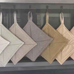 May include: A set of eight square quilted potholders hanging in a row, featuring a gradient of colors from off-white to light gray, tan, and soft pink. Each potholder has a small fabric loop at the top corner and a subtle star pattern throughout.
