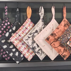May include: A collection of ten square fabric potholders hanging in a row, each featuring a unique pattern including ghosts, pumpkins, floral prints, and geometric designs in various colors like black, orange, white, and pink with fabric loops.
