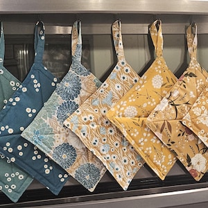 Puede incluir: Siete agarraderas de tela cuadradas colgadas en fila, con varios estampados florales en tonos azul verdoso, azul, amarillo mostaza y crema. Cada agarradera acolchada tiene un lazo de tela en la esquina superior para colgarla.