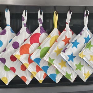 May include: A row of eight square fabric potholders hanging from hooks. The first four feature colourful polka dot patterns in blue, purple, orange, and green. The remaining four display vibrant star prints in yellow, orange, blue, and green.