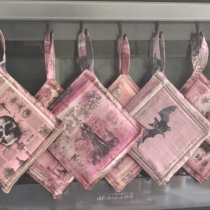 Op de afbeelding: Een set van acht vierkante stoffen pannenlappen die op een rij hangen, elk met een unieke roze en witte print met gotische invloeden. De ontwerpen bevatten een menselijke schedel, een figuur in een mantel, een vliegende vleermuis en een oog.