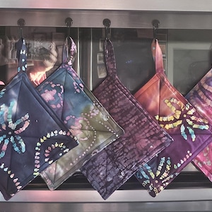 May include: A row of seven square fabric pot holders hanging from hooks. Each pot holder features a unique tie-dye or batik-style pattern in various colors including yellow, dark blue, purple, orange, and pink, all with a quilted diamond stitch.