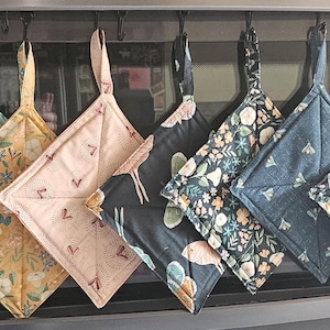 May include: Seven square fabric pot holders hanging in a row, each featuring a unique pattern including floral, botanical, and moth designs in various colors such as light blue, yellow, soft pink, dark gray, navy blue, and dark green.