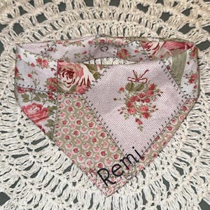 Peut inclure: Un bandana triangulaire pour animal avec un motif patchwork composé de divers imprimés floraux dans des tons rose, vert et blanc. Le tissu présente des roses et des petits pois. Le nom Remi est brodé en écriture noire sur la partie basse.
