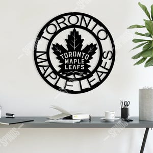 Könnte beinhalten: Schwarze Metall-Wanddekoration mit den Worten "Toronto Maple Leafs" um ein Ahornblatt-Emblem. Das kreisförmige Schild ist an einer weißen Wand über einem Schreibtisch angebracht. Es ist ein dekorativer Artikel.