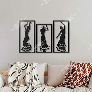 Könnte beinhalten: Set aus drei schwarzen Metall-Wandkunstwerken, die Flamenco-Tänzerinnen als Silhouette darstellen. Jede Tänzerin ist in einem rechteckigen schwarzen Rahmen eingefasst. Das Kunstwerk wird über einem Holzfuton mit Kissen und einem gemusterten Überwurf gezeigt. Der Text "SPINOS WALL DECOR" befindet sich unten.