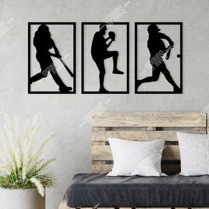 Puede incluir: Tres siluetas negras de metal de jugadores de béisbol en diferentes poses montadas en una pared gris claro. Las siluetas están enmarcadas en negro y están dispuestas en fila. La pared está detrás de un sofá de palets de madera con un cojín negro y almohadas blancas.