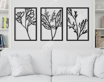 May include: A set of three rectangular black metal wall art panels featuring cut-out botanical designs of flowers, leaves, and branches. Each panel is framed in a thin black border and mounted on a plain wall above a white sofa.