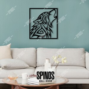 May include: Black metal wall decor featuring a stylized wolf howling at the moon. The wolf is in a square frame against a light blue wall. The decor is displayed on a white couch with a coffee table and a vase of white flowers.