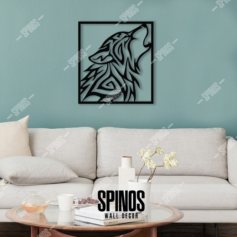 May include: Black metal wall decor featuring a stylized wolf howling at the moon. The wolf is in a square frame against a light blue wall. The decor is displayed on a white couch with a coffee table and a vase of white flowers.