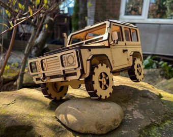 Puede incluir: Un modelo de madera de un Land Rover Defender marrón, un vehículo todoterreno popular. El modelo está detallado con una parrilla, faros y ruedas. Está sentado en una roca.