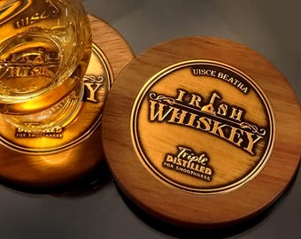 May include: Wooden coasters with a gold-colored emblem that reads "Irish Whiskey" and "Triple Distilled for Smoothness." A glass of whiskey sits on one coaster. The coasters are round and made of wood.