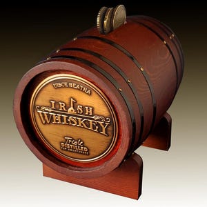 Può includere: Una salvadanaio a forma di botte in legno con un fronte color ottone. La banca presenta le parole "Irish Whiskey" e "Triple Distilled for Smoothness" sul davanti. Diverse monete sono visibili nella fessura superiore. La botte è sostenuta da un supporto in legno.