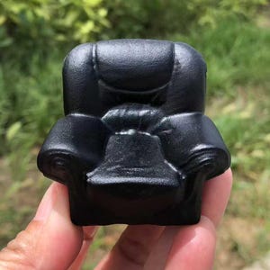 Pode incluir: Uma pequena poltrona preta em miniatura com almofadas e apoios de braços detalhados. A pequena cadeira é segurada em uma mão contra um fundo verde desfocado, sugerindo um ambiente externo. A cadeira é de cor escura sólida.