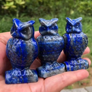 May include: Three carved lapis lazuli owl figurines. Each owl is a deep blue color with lighter gray and gold flecks. The owls have detailed features and are perched on a rectangular base. The background is blurred green foliage.