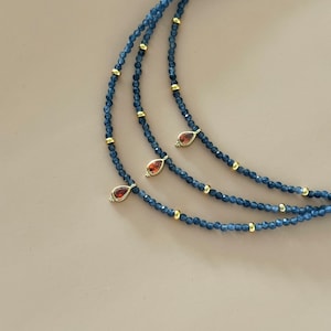 May include: A layered necklace with three strands of dark blue beads and gold accents. Each strand has a teardrop-shaped pendant with a red gemstone.