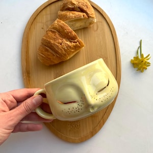 May include: A light yellow ceramic mug with a face design, featuring closed eyes and small black dots. Two croissants sit on a wooden oval serving board. A small bouquet of yellow flowers is also present.