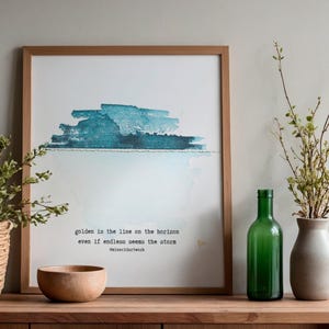 May include: A framed print with a blue and teal watercolour design and text. A green glass bottle, a beige vase, a wooden bowl, and a woven basket with greenery are on a wooden surface.
