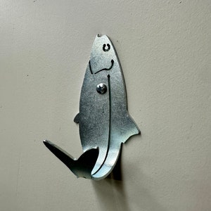 May include: A silver metal fish hook shaped like a fish with a smiling face. The hook is mounted on a light beige wall.