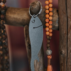 Puede incluir: Un llavero de metal gris con forma de pez con un abridor de botellas. El llavero cuelga de un anillo plateado. Un collar de cuentas naranjas y una estructura de madera están en el fondo. El pez tiene una boca sonriente y un ojo pequeño.