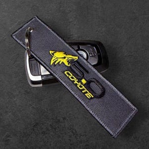 May include: A dark gray embroidered keychain with a yellow coyote logo and the text "5.0 Coyote". The keychain is attached to a silver key ring and rests on top of a car key.