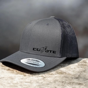 May include: A gray snapback hat with a black mesh back. The hat features the word "COYOTE" in black block letters with a coyote head graphic. The hat has a curved brim and a snap closure. The tag reads "THE AUTHENTIC SNAPBACK YUPOONG SINCE 1974."