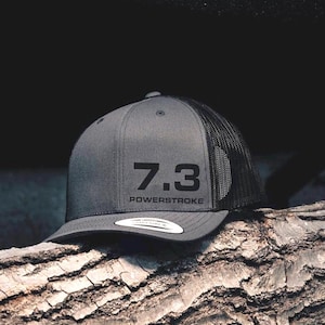 May include: A gray and black trucker hat with a black mesh back. The front of the hat has the text "7.3 POWERSTROKE" in black.