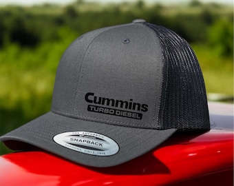 Può includere: Un cappello da camionista grigio e nero con un logo ricamato nero che dice "Cummins Turbo Diesel". Il cappello ha una parte posteriore in rete nera e una parte anteriore grigia con un adesivo argentato che dice "The Authentic Snapback - Founded Since 1974".